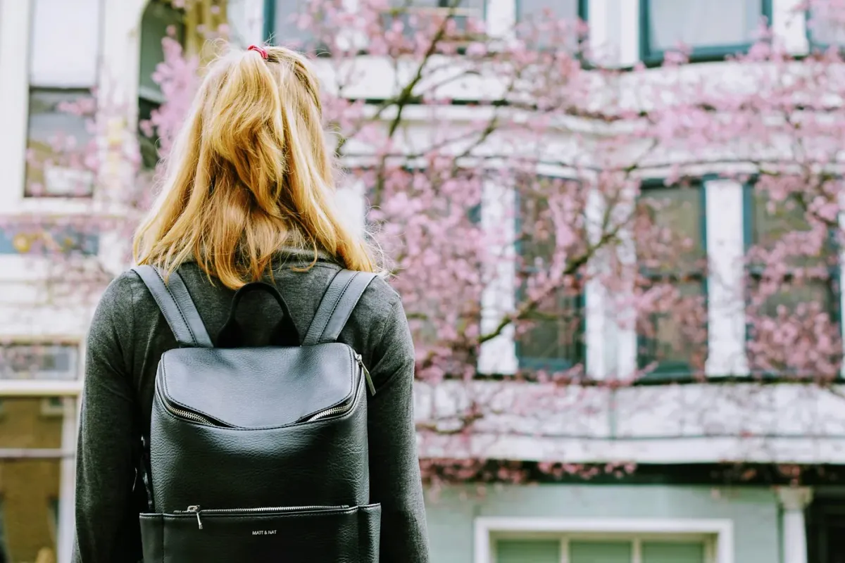 A blonde woman wearing a dark sweater and a leather backpack stands, facing away from the camera, looking toward an out-of-focus building and blooming tree with pink blossoms.