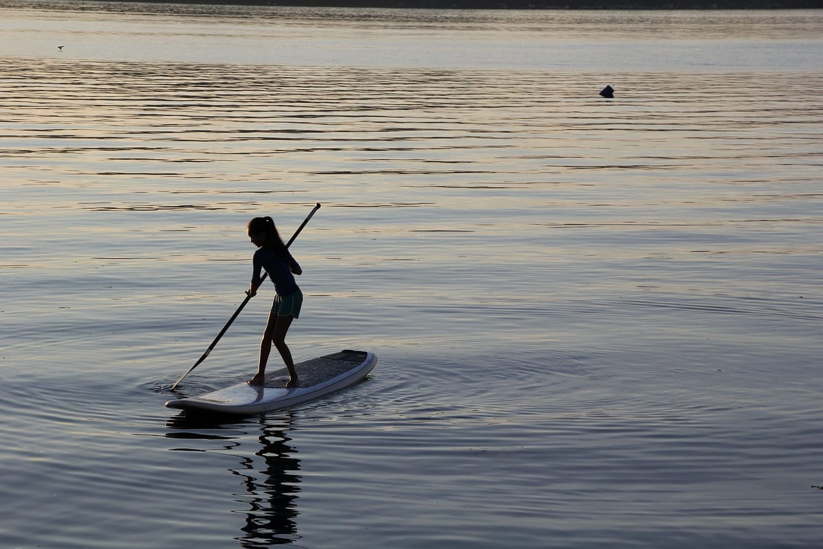 A young woman stands with an oar on a paddle board. She is surrounded by calm water reflecting the sunset.