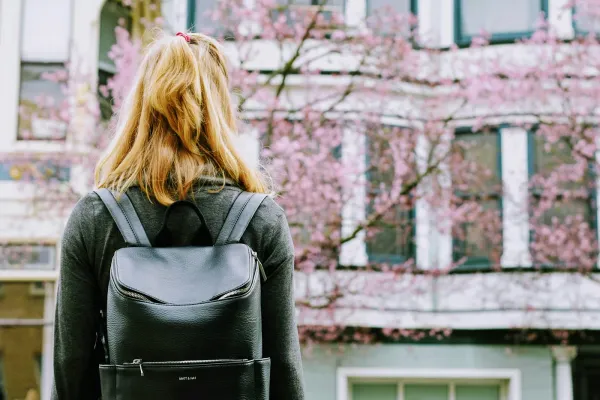 A blonde woman wearing a dark sweater and a leather backpack stands, facing away from the camera, looking toward an out-of-focus building and blooming tree with pink blossoms.