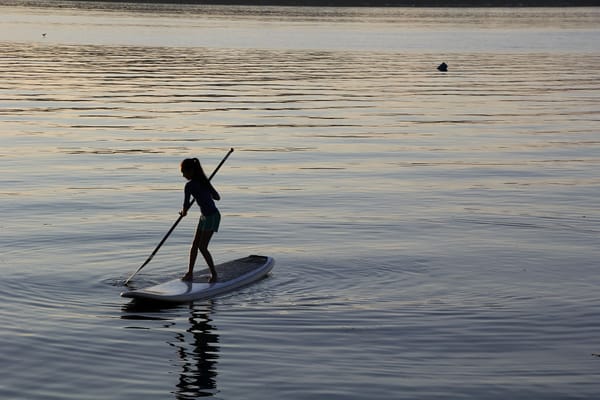 A young woman stands with an oar on a paddle board. She is surrounded by calm water reflecting the sunset.